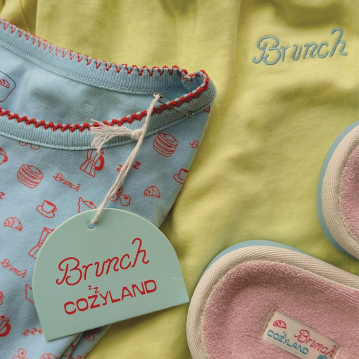 Close up of French Blue Cozyland PJ set showing red zigzag trim, breakfast print, Brunch hang tag, and pink terry slippers