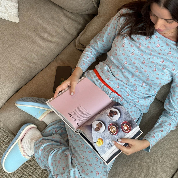 Woman reading on a beige couch wearing a French blue patterned pajama set with red waistband and light blue slippers