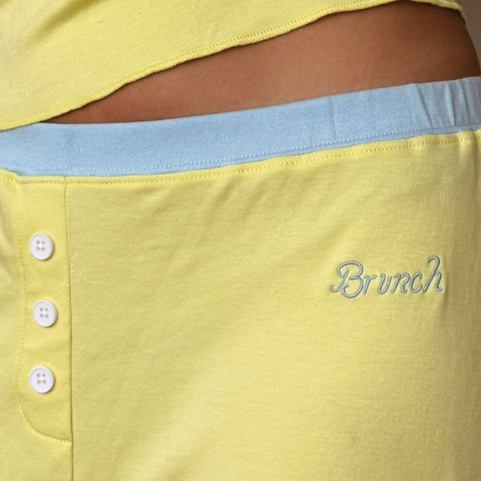 Close-up of pajama shorts showing light blue elastic waistband, three white corozo buttons, and Brunch script embroidery on sunshine yellow cotton