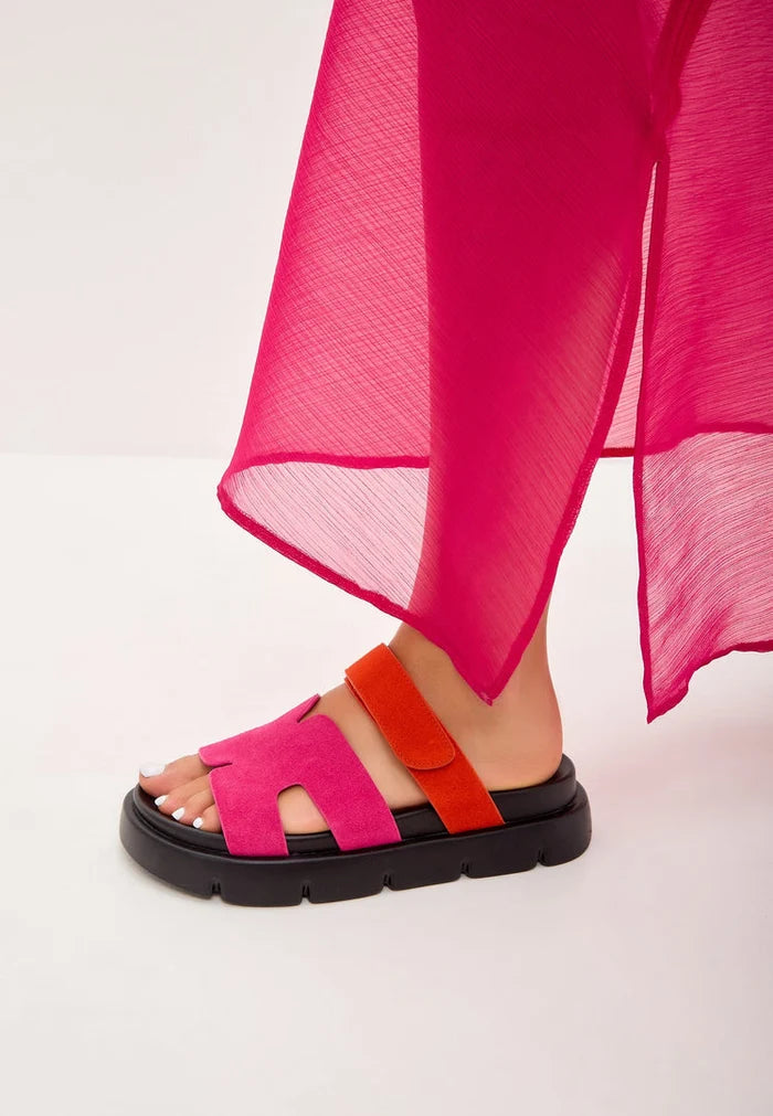 Close-up of a stylish foot wearing pink platform mules, partially covered by a flowing pink dress.