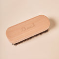 Top view of suede brush showing engraved Brunch wooden handle and dense horsehair bristles on a pale beige background