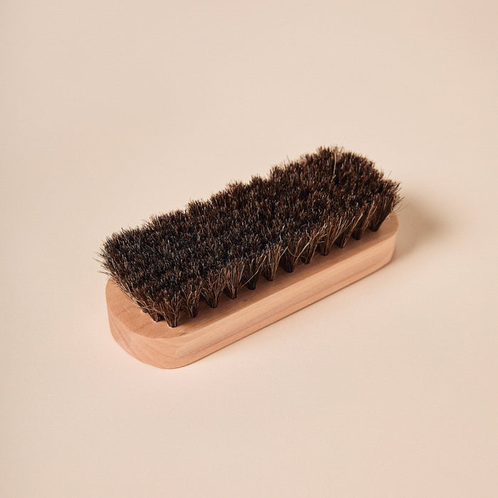 Angled top view of suede brush showing dense dark horsehair bristles and light wood handle on a pale beige background