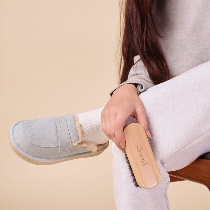 Hand holding a wooden suede brush brushing a light gray shearling-lined slipper on a seated model